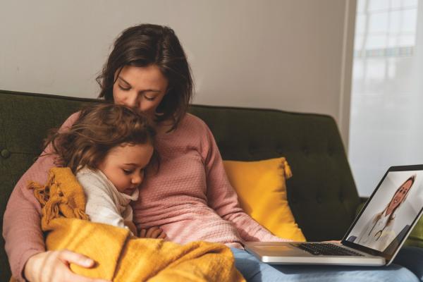 Mutter tröstet ihre kleine Tochter, während sie auf dem Sofa sitzt; auf dem Laptop ist eine Ärztin/ein Arzt zu sehen.